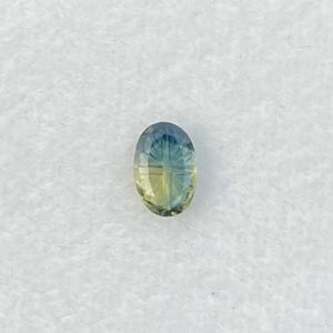 May include: An oval-cut gemstone featuring a color gradient that transitions from a deep blue at the top to a pale yellow at the bottom. The stone has a faceted surface with a symmetrical pattern of lines radiating from the center point.