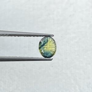 May include: An oval-shaped, faceted gemstone featuring a distinct cross pattern in the center. The stone displays a gradient of colors, transitioning from deep blue at the edges to a bright yellow hue in the middle, held by metal tweezers.
