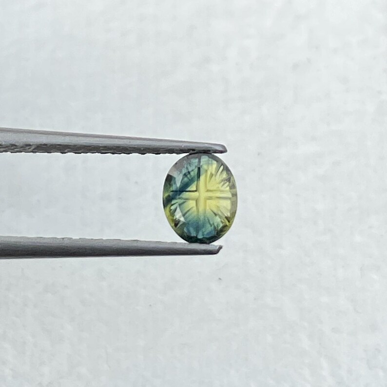 May include: An oval-shaped, faceted gemstone featuring a distinct cross pattern in the center. The stone displays a gradient of colors, transitioning from deep blue at the edges to a bright yellow hue in the middle, held by metal tweezers.