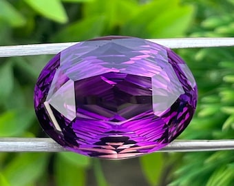 May include: A faceted, oval-shaped amethyst gemstone. The gem displays a deep purple hue with lighter shades and facets that catch the light. The gemstone is held by a silver-colored metal wire against a blurred green background.