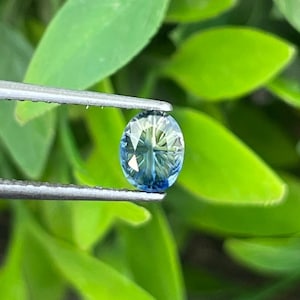 May include: An oval-cut gemstone with a faceted surface displaying a gradient of blue and yellow hues. The stone is held by a pair of silver-toned metal tweezers, showcasing its transparent quality and intricate internal light reflections.