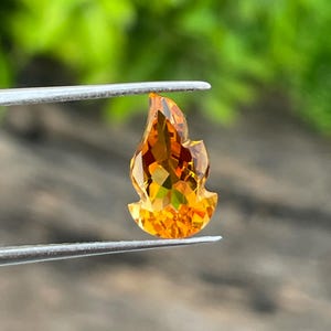 Könnte beinhalten: Ein facettierter, flammenförmiger Edelstein, der von einer Pinzette gehalten wird. Der Edelstein zeigt eine Reihe warmer Farben, von goldgelb bis tieforange, mit einer brillanten, reflektierenden Oberfläche. Der Schliff erzeugt mehrere Facetten.