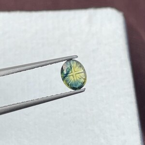 May include: An oval-shaped gemstone featuring a distinct cross pattern carved into its surface. The stone displays a gradient of colors, transitioning from a deep teal blue at the edges to a bright yellow hue in the center, held by metal tweezers.