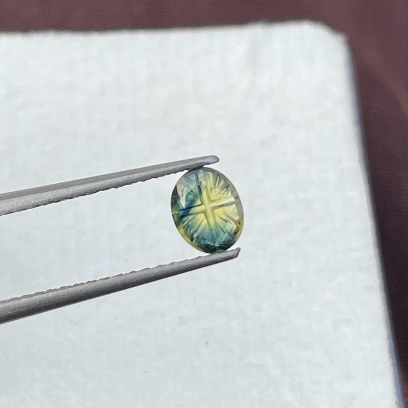 May include: An oval-shaped gemstone featuring a distinct cross pattern carved into its surface. The stone displays a gradient of colors, transitioning from a deep teal blue at the edges to a bright yellow hue in the center, held by metal tweezers.