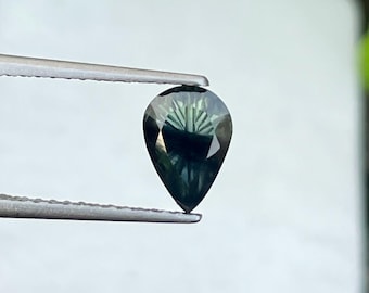 Puede incluir: Una piedra preciosa en forma de pera con una paleta de colores verde azulado profundo y verde oscuro. La piedra presenta una superficie facetada que crea un patrón radial de reflejos claros y oscuros en su interior, sujeta por pinzas.