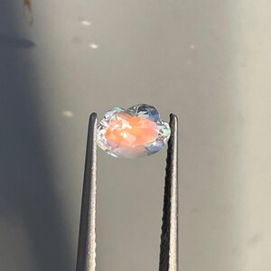Pode incluir: Uma pedra preciosa facetada de formato oval segurada por uma pinça de metal. A pedra é translúcida com uma borda externa clara e um centro vibrante de cor pêssego que reflete a luz com um brilho suave e iridescente na superfície.