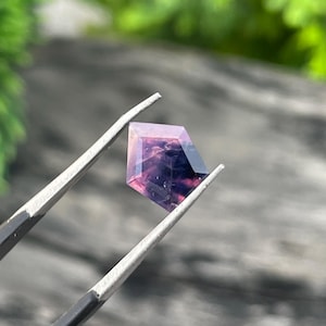 Puede incluir: Una gema facetada sostenida por unas pinzas. La gema tiene una forma geométrica con un degradado de colores, que va del rosa claro al morado oscuro. El fondo está borroso, con una superficie de madera y follaje verde.