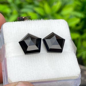 May include: Two loose, black, pentagon-shaped gemstones with a faceted surface. The stones are displayed side-by-side on a white, textured foam insert inside a clear plastic container, showcasing their geometric cut and dark, reflective finish.