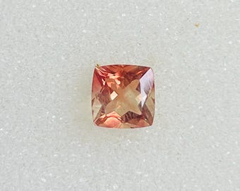 May include: A square-cut gemstone with a peach and light brown color gradient. The gemstone has a faceted surface that reflects light, creating a sparkling effect. The gem is set against a white background.