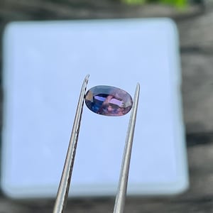 May include: An oval-shaped gemstone held by tweezers. The gem displays a blend of purple, blue, and pink hues. The background is a blurred white surface and a wooden texture.