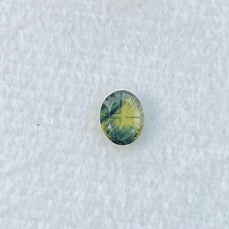 May include: An oval-cut gemstone featuring a gradient of colors transitioning from a deep forest green at the edges to a bright, translucent yellow at the center, with a faceted surface that creates a distinct cross-like pattern of light reflection.