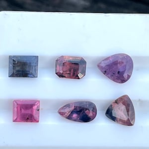 May include: Assortment of six gemstones in various shapes: square, emerald cut, and pear-shaped. The gems display colors of pink, purple, and blue. The gemstones are set against a white background.