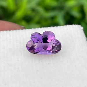 Peut inclure: Une pierre précieuse améthyste violette à facettes taillée en forme de nuage. La pierre présente de multiples facettes géométriques qui reflètent la lumière, créant des nuances variées de violet profond et de lavande sur sa surface polie.