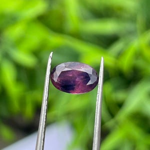 Op de afbeelding: Een ovaal geslepen edelsteen met een dieppaarse kleur, vastgehouden door een zilveren pincet. De facetten van de edelsteen reflecteren het licht, waardoor de helderheid en kleur zichtbaar worden. De achtergrond is zachtgroen.