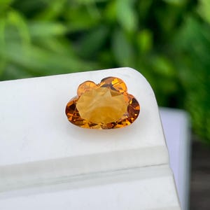 Può includere: Una gemma giallo oro sfaccettata, tagliata a forma di nuvola. La pietra presenta una superficie brillante a facce multiple che riflette la luce, mostrando una calda tonalità ambrata traslucida con una finitura lucida.