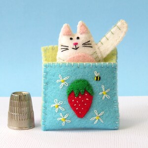 Puede incluir: Un gato de fieltro blanco con mejillas rosadas y una nariz negra asoma de una caja de fieltro azul con un bordado de fresa y abeja. La caja est&aacute; rodeada de margaritas blancas.