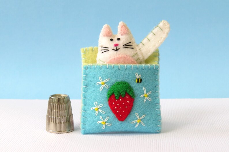 Puede incluir: Un gato de fieltro blanco con mejillas rosadas y una nariz negra asoma de una caja de fieltro azul con un bordado de fresa y abeja. La caja est&aacute; rodeada de margaritas blancas.