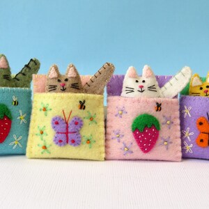 Puede incluir: Cuatro bolsillos de fieltro con forma de gato con detalles bordados. Cada bolsillo presenta un color y un dise&ntilde;o diferente, incluyendo una mariposa, una fresa y una abeille.