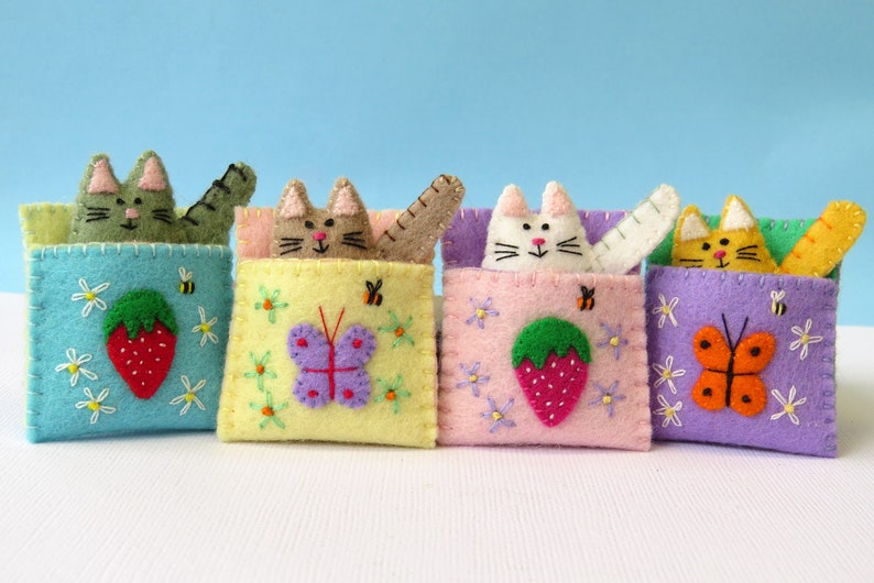 Puede incluir: Cuatro bolsillos de fieltro con forma de gato con detalles bordados. Cada bolsillo presenta un color y un dise&ntilde;o diferente, incluyendo una mariposa, una fresa y una abeille.