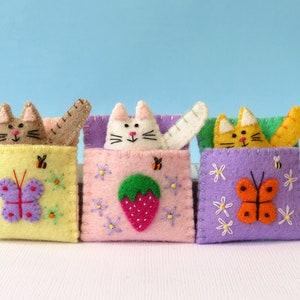 Puede incluir: Tres juguetes de fieltro con forma de gato, cada uno con un color y dise&ntilde;o diferente. El primer gato es marr&oacute;n con un bolsillo amarillo y una mariposa morada. El segundo gato es blanco con un bolsillo rosa y una fresa verde. El tercer gato es naranja con un bolsillo morado y una mariposa naranja.