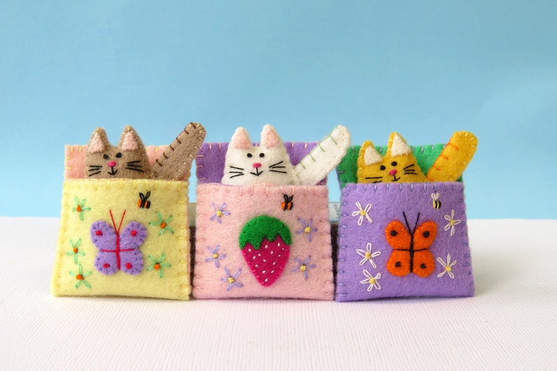 Puede incluir: Tres juguetes de fieltro con forma de gato, cada uno con un color y dise&ntilde;o diferente. El primer gato es marr&oacute;n con un bolsillo amarillo y una mariposa morada. El segundo gato es blanco con un bolsillo rosa y una fresa verde. El tercer gato es naranja con un bolsillo morado y una mariposa naranja.