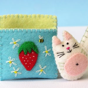 Puede incluir: Un gato de fieltro con la barriga rosa y la cara blanca, sosteniendo una bolsa de fieltro azul con un bordado de fresa y margarita.
