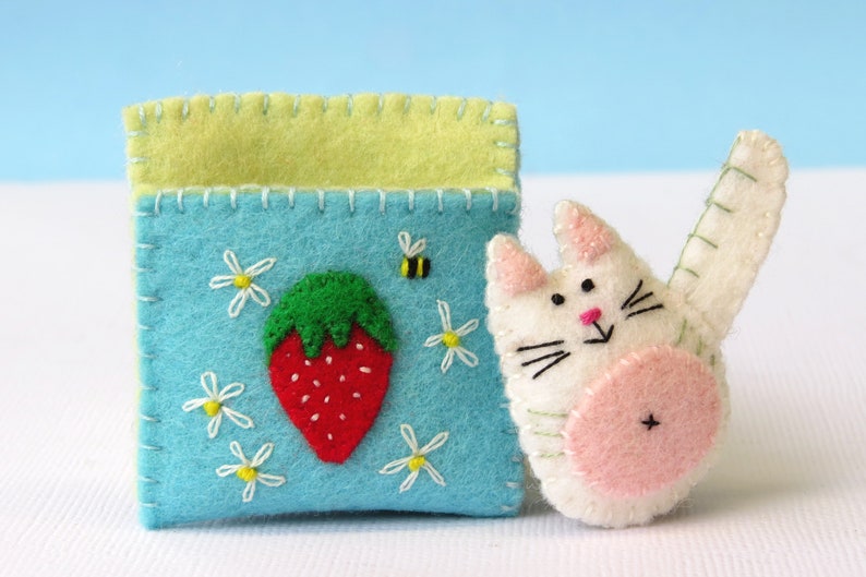 Puede incluir: Un gato de fieltro con la barriga rosa y la cara blanca, sosteniendo una bolsa de fieltro azul con un bordado de fresa y margarita.
