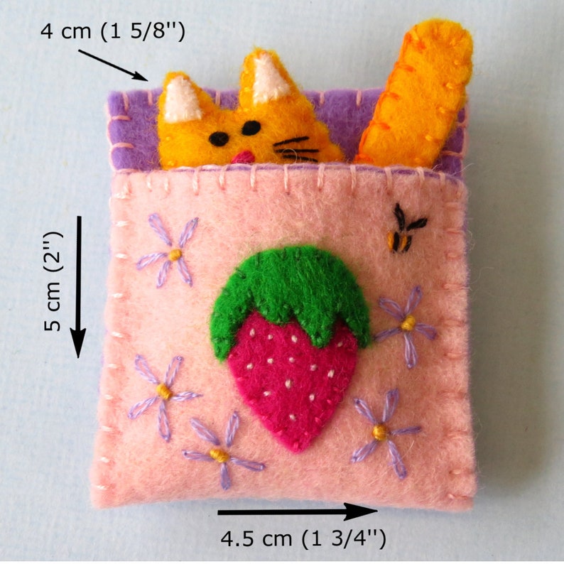Puede incluir: Una bolsa de fieltro rosa con un dise&ntilde;o de fresa y un peque&ntilde;o gato naranja asom&aacute;ndose por la parte superior. La bolsa mide 10 cm de ancho, 11,5 cm de largo y 5 cm de alto.