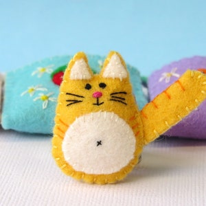 Puede incluir: Un gato de fieltro amarillo con vientre blanco y nariz rosa. El gato tiene una cola larga y est&aacute; sentado sobre una superficie blanca.