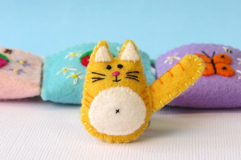Puede incluir: Un gato de fieltro amarillo con vientre blanco y nariz rosa. El gato tiene una cola larga y est&aacute; sentado sobre una superficie blanca.