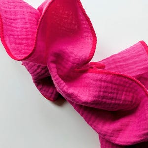 Könnte beinhalten: Ein leuchtend fuchsiafarbenes Musselintuch mit einer geknitterten, strukturierten Stoffoberfläche und einem passenden, leuchtend pinkfarbenen Saum. Das leichte Textil ist gefaltet und drapiert, um das weiche, mehrlagige und atmungsaktive Gewebe zu zeigen.