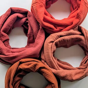 Könnte beinhalten: Eine Sammlung von fünf Schlauchschals in Rot- und Orangetönen. Die Schals sind aus einem strukturierten Stoff gefertigt und kreisförmig angeordnet. Die Farben reichen von einem tiefen Ziegelrot bis zu einem leuchtenden Orange.