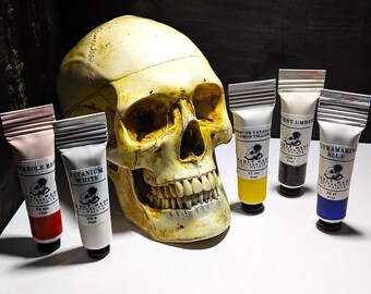 May include: A human skull sits in front of six tubes of oil paint. The tubes are labeled with the paint colors:  Titanium White,  Pyrrole Red,  Burnt Umber,  Ultramarine Blue,  and  Cadmium Lemon Yellow.  The paint tubes are from the brand "Badly Made Professional Oil Paints."