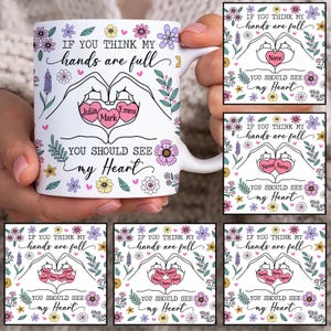 May include: White ceramic mug with a floral design and the text "If you think my hands are full, you should see my heart". The mug features a heart shape formed by two hands with the names "Julia", "Mark", and "Emma" inside the heart.