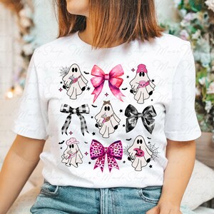 Op de afbeelding: Wit T-shirt met een roze, zwart en wit spook- en strikdesign. De spoken dragen hoeden en houden verschillende voorwerpen vast, waaronder een pompoen, een spinnenweb en een kopje koffie. De strikken zijn in verschillende patronen, waaronder ruit, stippen en effen kleuren.