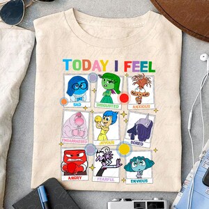 May include: A beige t-shirt with the words 'TODAY I FEEL' printed on the front. Below the words are nine squares with different characters from the movie Inside Out. Each square has a different emotion written below the character. The emotions are: Sad, Disgusted, Anxious, Embarrassed, Joyous, Bored, Angry, Fearful, and Envious.