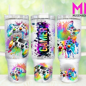 May include: A white tumbler with a handle features a colorful design of video game controllers and paint splatters. The text "GAMER" is printed on the tumbler.