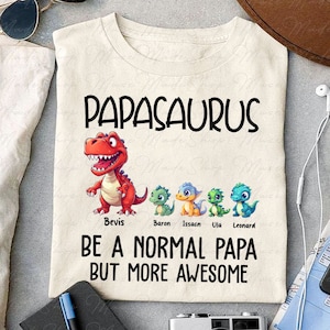 May include: A beige t-shirt with the text "PAPASAURUS" in black. Below the text is a red dinosaur with four smaller dinosaurs in a line below it. The text "Bevis, Baron, Issacn, Ula, Leonard" is below the dinosaurs. The text "BE A NORMAL PAPA BUT MORE AWESOME" is below the dinosaurs.
