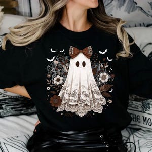 Puede incluir: Camiseta negra con un fantasma blanco que lleva un lazo marr&oacute;n y encaje. El fantasma est&aacute; rodeado de flores marrones y telara&ntilde;as.