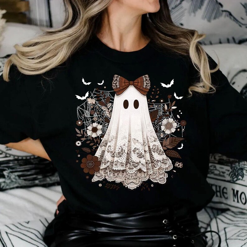 Puede incluir: Camiseta negra con un fantasma blanco que lleva un lazo marr&oacute;n y encaje. El fantasma est&aacute; rodeado de flores marrones y telara&ntilde;as.