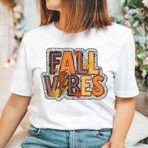 May include: White t-shirt with a graphic design that says "Fall Vibes" in orange, brown, and black letters with a leopard print, lightning bolt, and fall leaves.