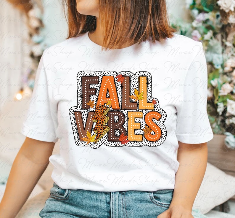 May include: White t-shirt with a graphic design that says "Fall Vibes" in orange, brown, and black letters with a leopard print, lightning bolt, and fall leaves.