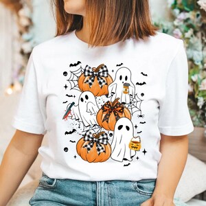 May include: White t-shirt with a Halloween design featuring ghosts and pumpkins. The ghosts are wearing hats and holding coffee cups and candy buckets. The pumpkins have black and white checkered bows.