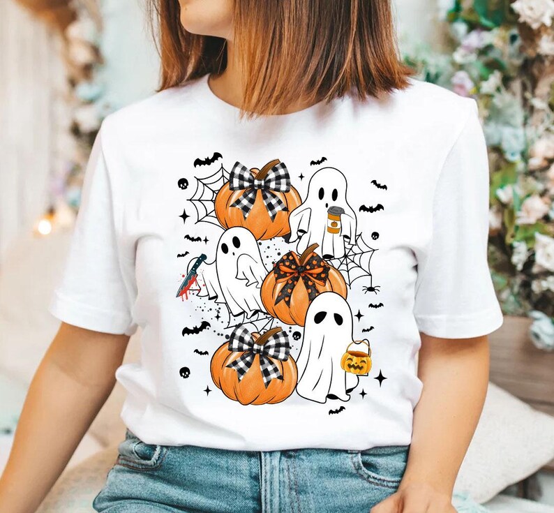 May include: White t-shirt with a Halloween design featuring ghosts and pumpkins. The ghosts are wearing hats and holding coffee cups and candy buckets. The pumpkins have black and white checkered bows.