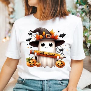 Puede incluir: Camiseta blanca con un fantasma de dibujos animados que lleva un sombrero de bruja y sostiene un l&aacute;piz que dice "Mrs. Smith". El fantasma est&aacute; rodeado de murci&eacute;lagos, telara&ntilde;as y calabazas.