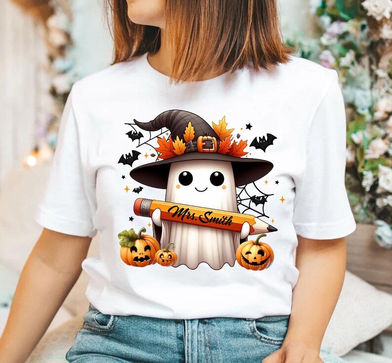 Puede incluir: Camiseta blanca con un fantasma de dibujos animados que lleva un sombrero de bruja y sostiene un l&aacute;piz que dice "Mrs. Smith". El fantasma est&aacute; rodeado de murci&eacute;lagos, telara&ntilde;as y calabazas.