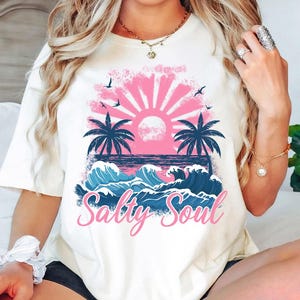 May include: White t-shirt featuring a graphic of a pink sunset over blue waves and palm trees. The text "Salty Soul" is printed in pink below the graphic.