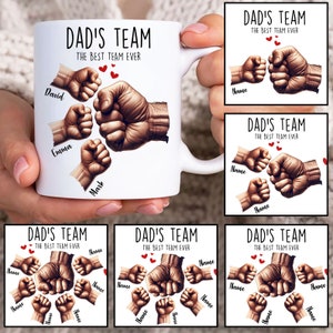 Op de afbeelding: Een witte keramische mok met een ontwerp van stotende vuisten. De tekst "DAD'S TEAM THE BEST TEAM EVER" staat op de mok gedrukt. De namen David, Emma en Mark staan op de mok gedrukt.