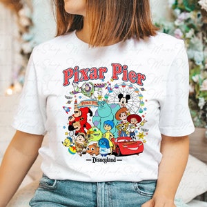 Puede incluir: Camiseta blanca con un gr&aacute;fico colorido de personajes de Pixar de diferentes pel&iacute;culas, incluyendo Buzz Lightyear, Woody, Mike Wazowski, Sulley y Rayo McQueen. El texto "Pixar Pier - Disneyland" est&aacute; impreso en la camiseta.