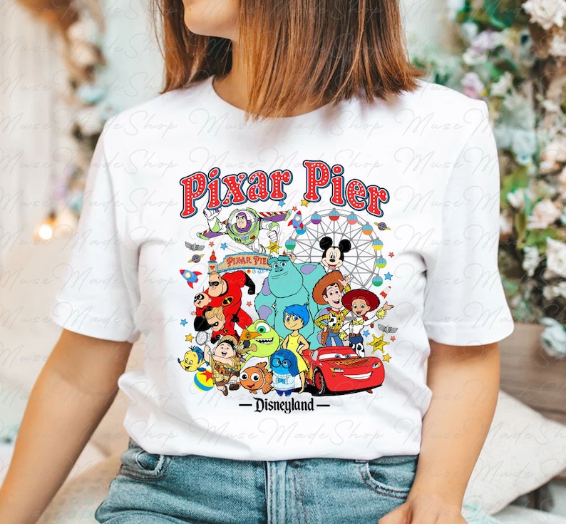 Puede incluir: Camiseta blanca con un gr&aacute;fico colorido de personajes de Pixar de diferentes pel&iacute;culas, incluyendo Buzz Lightyear, Woody, Mike Wazowski, Sulley y Rayo McQueen. El texto "Pixar Pier - Disneyland" est&aacute; impreso en la camiseta.